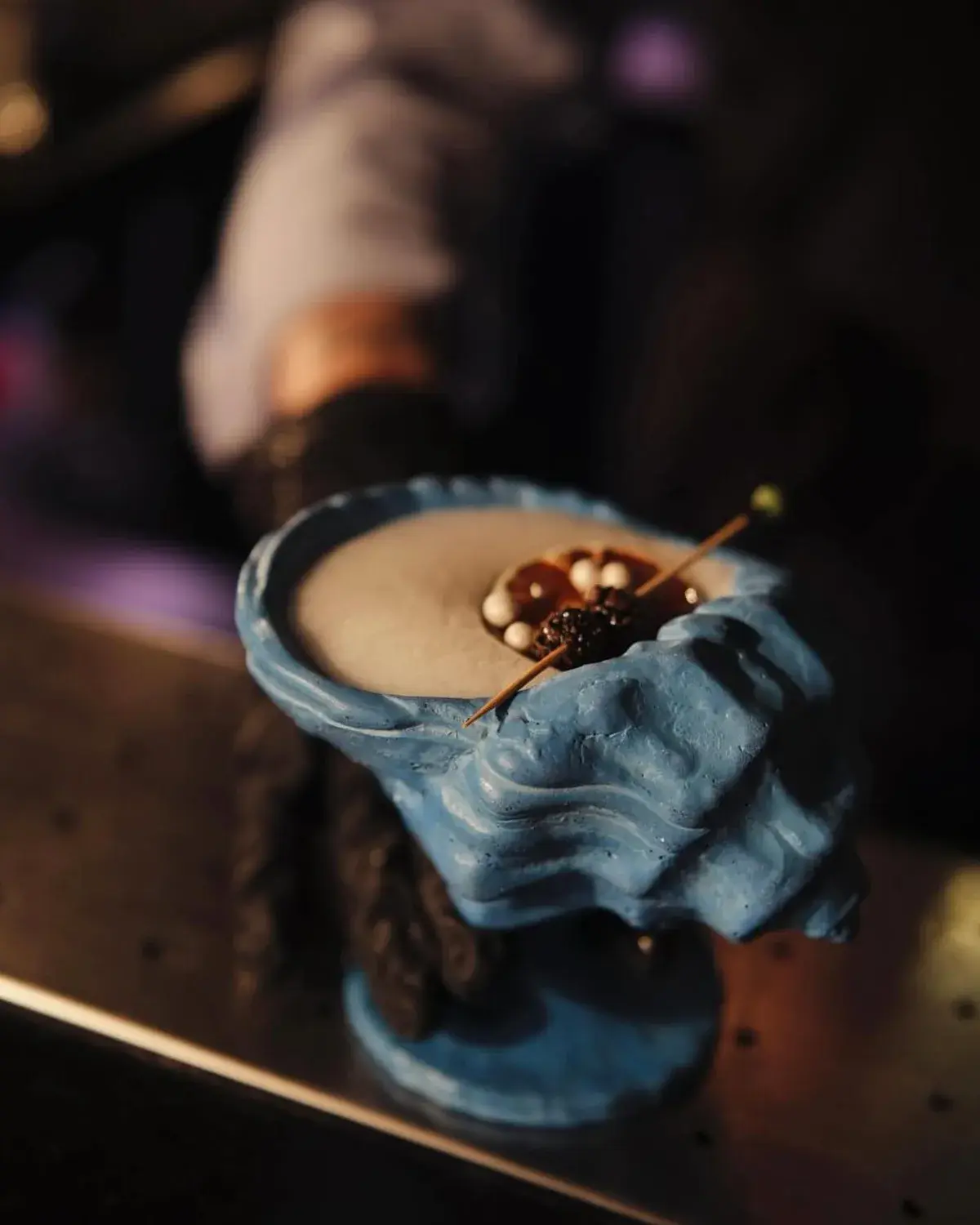 Blue shell-shaped concrete vessel holding a cocktail, held over a bar