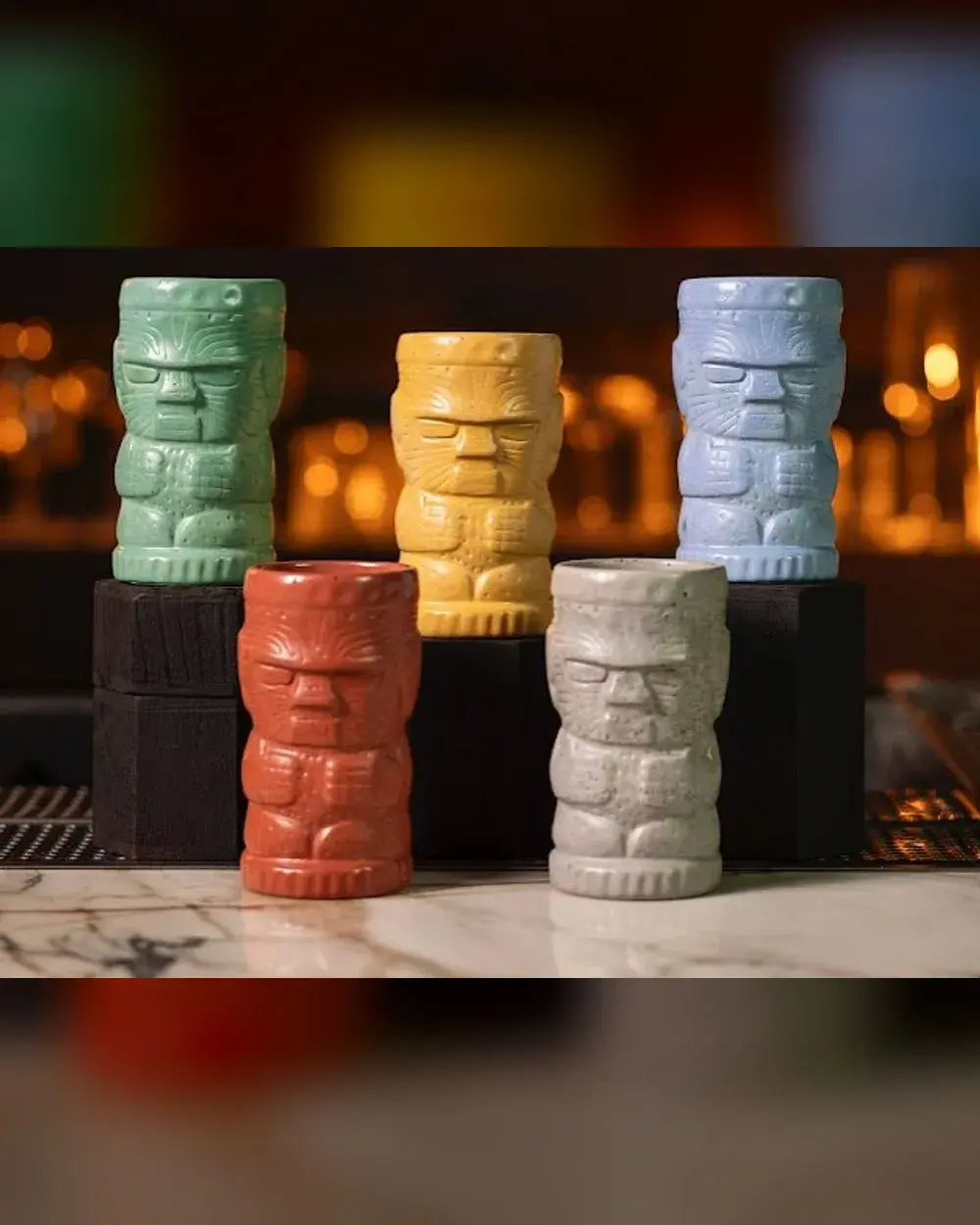 Five concrete tiki mugs of one shape in green, yellow, pale blue, red and grey, lined up on a bar