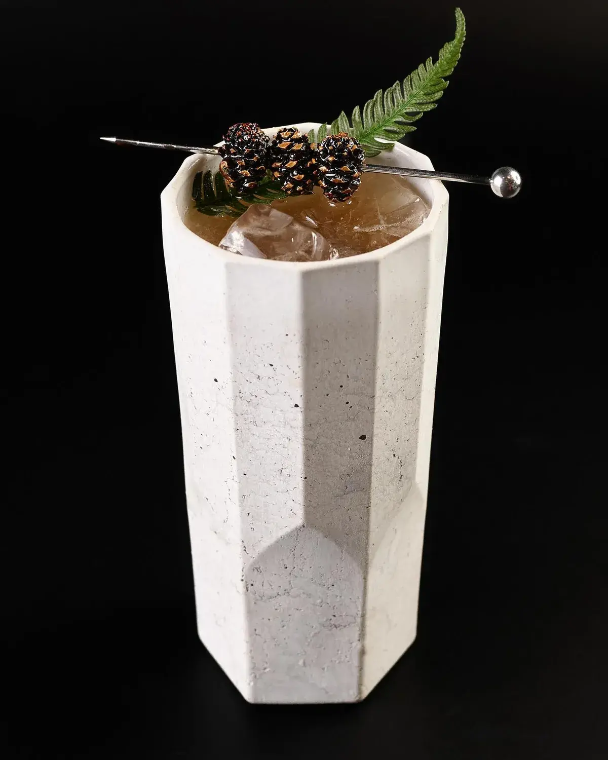Tall faceted concrete tumbler in white, served over ice with a fern garnish